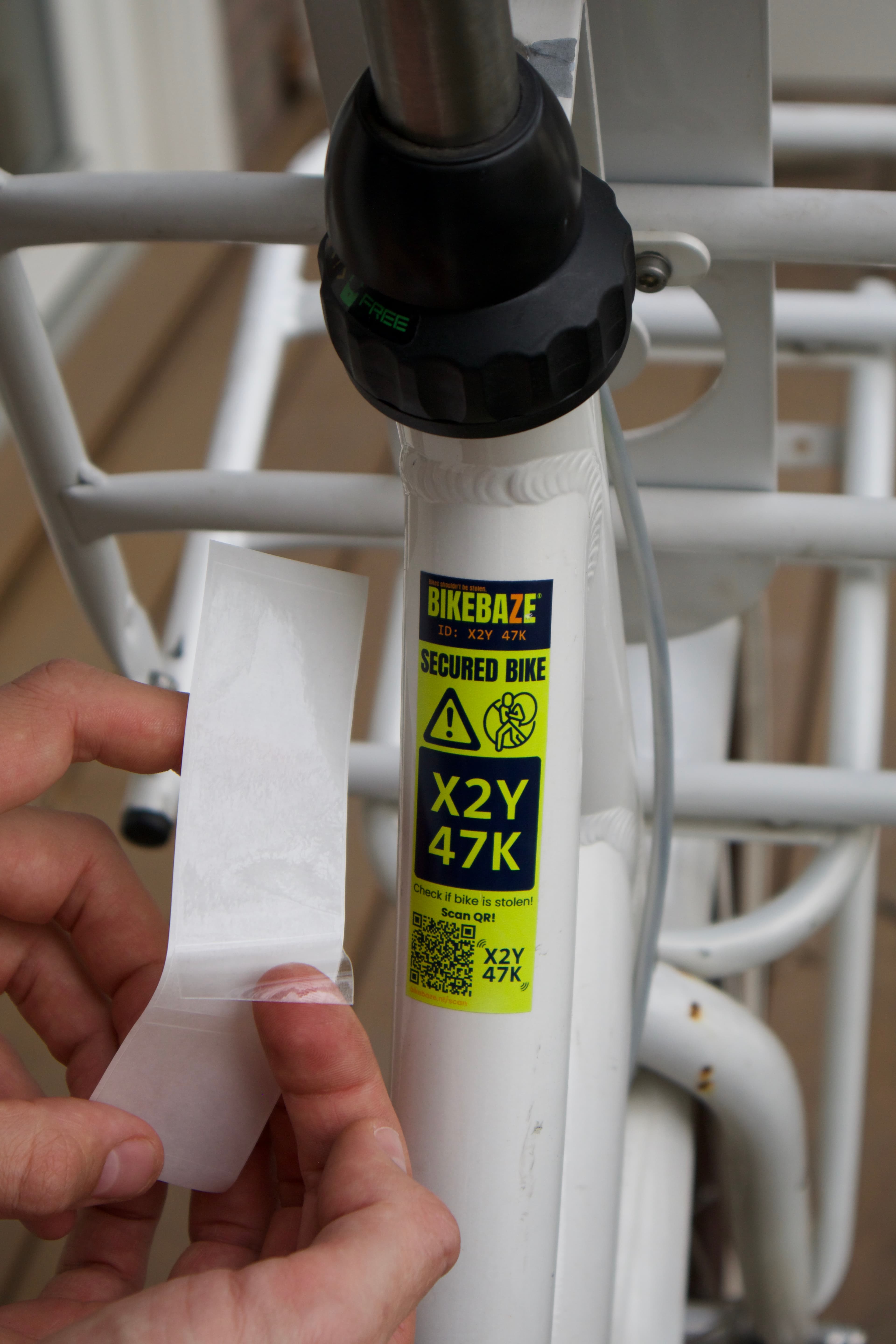 Hand brengt een transparante SafeTag Protect beschermlaag aan over een gele BIKEBAZE SafeTag op een wit fietsframe.